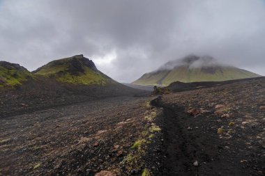 Bu çarpıcı görüntü, yemyeşil tepelerin bulutlu gökyüzünün altında kayalık siyah yollarla buluştuğu dramatik bir volkanik manzara yakalar ve büyüleyici ve gizemli bir ortam yaratır..