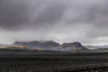 Karanlık volkanik toprağı, çarpan dağları ve dönen gri bulutları gösteren karamsar İzlanda manzarasının büyüleyici görüntüsü gerçeküstü ve büyüleyici bir atmosfer yaratıyor..