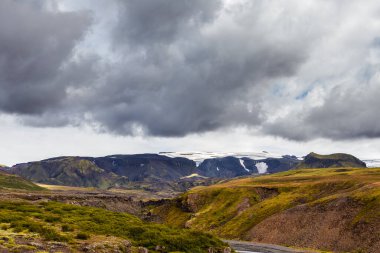 Bu görüntü İzlanda dağlarının güzelliğini yakalar, bulutlu bir gökyüzünün altında görkemli dağları ön planda kar kalıntıları ve yemyeşil vadilerle gösterir..