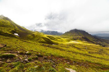 Bu resim, yuvarlanan yeşil tepeler, kar yığınları ve dağları örten dramatik bulutların yer aldığı nefes kesici bir manzara sunuyor. İzlanda 'nın sakin ortamını yansıtıyor..