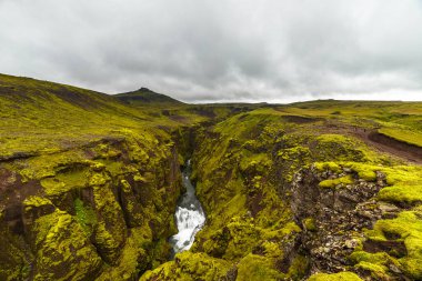 Bu büyüleyici görüntü, İzlanda 'da dramatik bulutlu gökyüzü altında yosun kaplı yosun kaplı yamaçların çerçevelediği, içinden akan bir nehrin aktığı yemyeşil bir kanyonu gözler önüne seriyor..