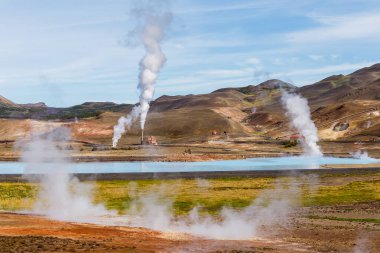 Bu çarpıcı görüntü İzlanda jeotermal manzarasını sergiliyor, buharı tüten bacalar, dingin bir su kütlesi, ve arka planda engebeli dağlar, doğanın güzelliğini yansıtıyor..
