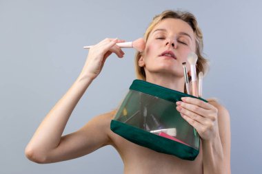 Bu büyüleyici görüntüde bir kadın makyaj yapıyor, çeşitli fırçalarla makyaj yapıyor ve elinde güzellik malzemeleriyle dolu yeşil bir kozmetik çanta tutuyor..