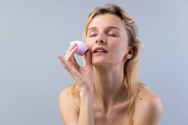 Bu resimde, dalgalı saçlı güzel bir kadın mor makyaj süngeri kullanarak makyaj yapıyor. Rahatlamış bir atmosfer sergiliyor ve rutin cildine bağlılığını gösteriyor.