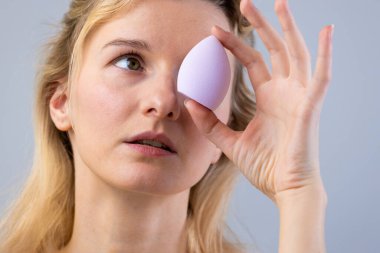 Bu resimde, bir kadın gözünün yanında mor bir güzellik blenderi tutarken düşünceli görünüyor, makyaj uygulamasını ve kusursuz kozmetik tekniklerini sembolize ediyor..