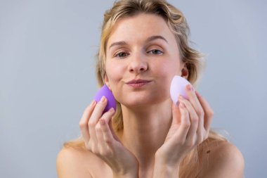 Açık renk saçlı genç bir kadın mor ve pembe bir süngeri elinde tutuyor, kendine güvenerek gülümsüyor. Görüntü, güzellik rutin sürecinde bir neşe ve yaratıcılık anı yakalar..