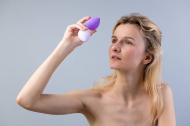 Görüntüde uzun saçlı bir kadın var, iki güzel harmanlanmış süngere özenle bakıyor, biri mor, biri beyaz, makyaj uygulama aletlerinin önemini vurgulayan..