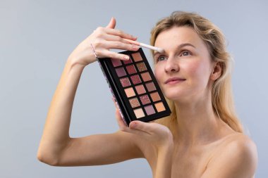 Güzel genç bir kadın renkli bir göz farı paleti kullanarak nazikçe göz makyajı yapıyor. Görüntü, canlı renkleri ve odaklanmış ifadesini vurguluyor..