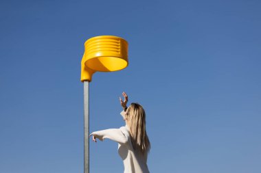 Parlak mavi gökyüzüne çarpan sarı bir enstalasyonla etkileşime giren bir kadın, sahnede canlı ve sanatsal bir atmosfer yaratıyor..