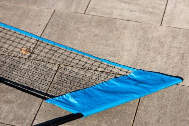 Beton bir yüzeyde duran parlak mavi bir tenis ağının yakın plan görüntüsü. Ağlar karmaşık bir şekilde örülmüş, güneşli bir açık havada spor malzemelerinin detaylarını gösteriyor..