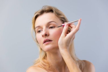 Odaklanmış bir kadın pembe saplı bir fırça kullanarak dikkatlice göz kalemi sürüyor. Güzelliği ve makyaj sanatını yumuşak gri bir arka plana yansıtarak kendini ifade etme sanatını sergiliyor..