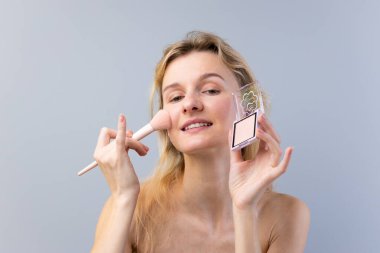 Bu görüntüde, yüzüne makyaj yapan, makyaj fırçası ve paletini sergiliyor, sakin, açık gri bir arkaplan üzerine kurulmuş, güzelliği ve kendine bakımı kutlayan neşeli bir kadın görülüyor..