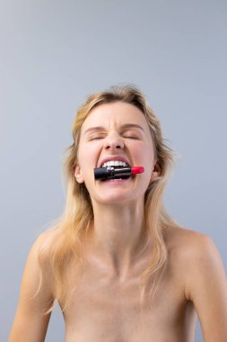 Bu resimde sarı saçlı neşeli bir genç kadın var. Ağzında bir tüp ruj tutuyor. Güzelliği, kendini ifade etme ve makyaj sanatını vurguluyor..