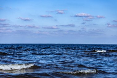 Bu sakin deniz manzarası, kıyı şeridiyle etkileşime geçen nazik dalgaları sergiliyor. Yumuşak bulutlarla benekli parlak mavi gökyüzü, huzur ve sükuneti davet ediyor..