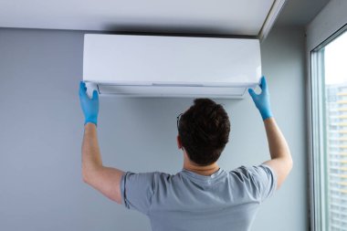 Mavi eldiven giyen bir kişi modern bir oturma odasına klima takıyor. Bu sahne uygun HVAC kurulumunun ev konforu ve verimliliği için önemini vurgular.