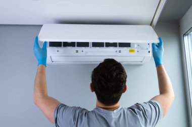 Bir teknisyen duvara monte edilmiş klima ünitesini ayarlarken, koruyucu eldivenler giyerken ve verimli soğutma işlemleri için modern bir iç mekanda düzgün bakım sağlarken görülüyor..