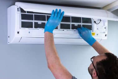 Mavi eldiven giyen bir HVAC teknisyeni, duvarlara monte edilmiş bir havalandırma ünitesinden filtreyi çıkarmak için özenle çalışıyor..