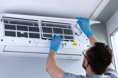 Bir havalandırma teknisyeni, hava kalitesini ve sistem performansını artırmak için filtreyi temizlemeye veya değiştirmeye odaklanan, duvarda kurulu bir klima ünitesi üzerinde çalışır..