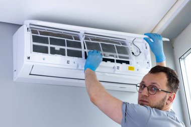 Eldiven giyen bir HVAC teknisyeni duvarda asılı bir havalandırma ünitesini dikkatlice inceliyor ve içeride rahat yaşayabilmesi için en uygun performans ve temizliği sağlıyor..