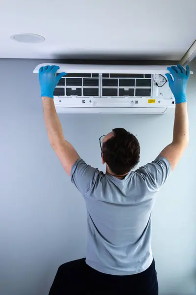 Mavi eldiven giyen bir teknisyen duvara monte edilmiş bir klima ünitesini dikkatlice tamir ediyor ve havalandırma tamir sırasında düzgün bakım ve güvenliğin önemini gösteriyor..