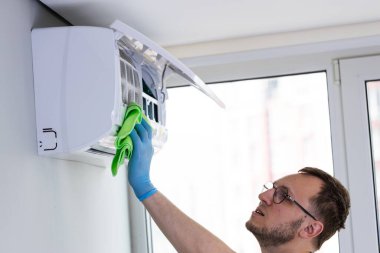 Görüntüde mavi eldivenli bir kişinin yeşil bir bez ile klimayı temizlediği, çağdaş bir yaşam alanında hijyen ve verimli hava akımını teşvik ettiği görülüyor..