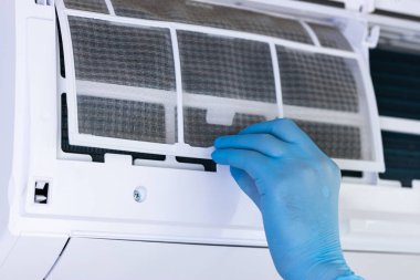 Mavi eldiven giyen bir elin yakın plan görüntüsü, havalandırma ünitesinden havanın kalitesini ve konforunu arttırmak amacıyla ustalıkla filtreyi çıkartıyor..