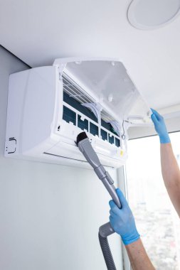 Eldiven giyen bir kişi vakum eklentisi kullanarak havalandırma ünitesini temizlerken görülüyor. Bu bakım basamağı en uygun işlevsel ve kapalı alandaki hava kalitesinin artırılması için hayati önem taşıyor..