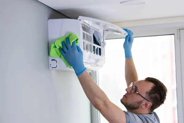 Kendini adamış bir HVAC teknisyeni, kapalı alanda koruyucu eldivenler giyerken yeşil bir mikrofiber bez kullanarak, duvarlara monte edilmiş bir klima ünitesinde temizlik yapıyor..