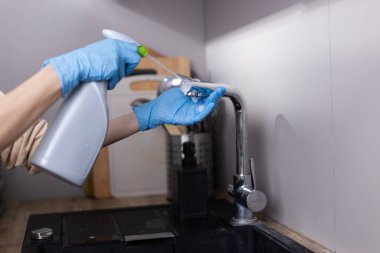 Mutfak musluğunu sprey şişeyle temizleyen birinin detaylı görüntüsü. Eller mavi eldivenlerle korunuyor, modern bir mutfak ortamında temizlik ve hijyen üzerine odaklanıyorlar..