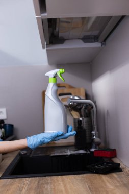 Görüntüde mavi eldivenli bir el, sprey bir şişe tutuyor, lavabo, temizlik aletleri ve cilalı bir tezgahın yer aldığı modern bir mutfak zeminine karşı kurulmuş..