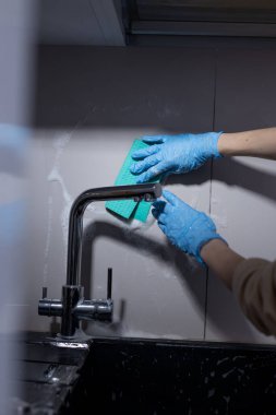 Bir birey, ev ortamlarında temizlik ve temizliğin önemini vurgulamak için mavi lastik eldiven giyerken canlı yeşil bir süngerle mutfak musluğunu temizlemekle meşgul..