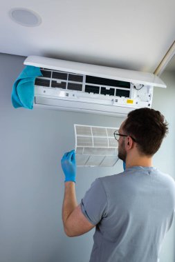 Bir kişi, eldiven giyerken havalandırma ünitesinin filtresini temizlerken, modern bir ortamda iyi hava akışı ve hijyeni sağlarken, ev bakımı çalışmaları sergilerken görülüyor..