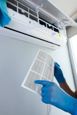 Görüntü, mavi lateks eldiven giyen, havalandırma filtresini dikkatlice inceleyen, düzenli temizliğin ev konforu ve hava kalitesi açısından önemini vurgulayan birini gösteriyor..