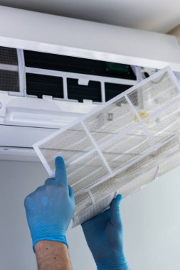 Mavi eldiven giyen bir kişi, temiz hava kalitesini, performansını ve soğutma ünitesinin ömrünü uzatmak için kirli klima filtresini dikkatlice çıkarır..
