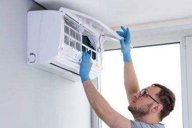 Bir teknisyen, duvarlara monte edilmiş bir havalandırma ünitesi üzerinde özenle çalışıyor, teftiş ve bakım görevlerine odaklanıyor. Profesyonel güvenlik için koruyucu eldiven giyer..