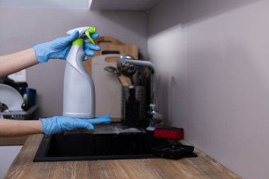 Mavi lateks eldiven giyen bir kişi, çağdaş bir mutfakta gri bir temizlik spreyi tutuyor. Tezgahları ve bulaşıkları etkin bir şekilde temizlemeye hazırlanırken temizliği vurguluyor..