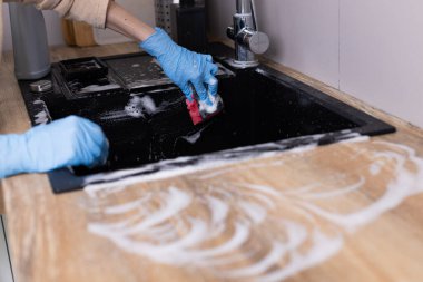 Görüntü, mavi eldiven giymiş bir kişiyi renkli bir süngerle lavaboyu fırçalarken yakalıyor. Mutfaktaki temizliğin önemini vurguluyor ve iyi bir hijyen sağlıyor..