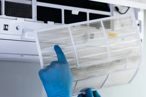 Mavi eldiven giyen bir şahıs, havalandırma ünitesinden kirli hava filtresini çıkartıyor ve kapalı hava kalitesini artırmak için düzenli bakım gereğini vurguluyor..