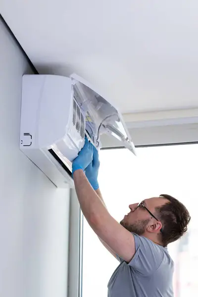 Bir teknisyen, duvarlara monte edilmiş bir klimayı dikkatlice tamir ediyor, eldiven takıyor ve iyi ışıklandırılmış bir iç mekanda bakım görevlerine odaklanıyor..