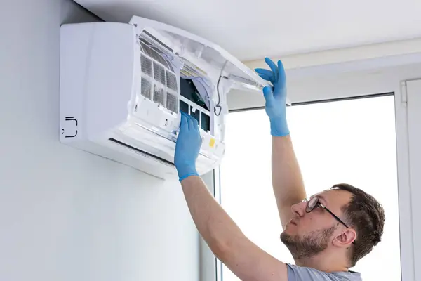 Bir teknisyen, duvarlara monte edilmiş bir havalandırma ünitesi üzerinde özenle çalışıyor, teftiş ve bakım görevlerine odaklanıyor. Profesyonel güvenlik için koruyucu eldiven giyer..