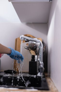 Bu resim, eldivenli birinin musluğu çalıştırdığı anı yakalar, sabunlu suyun akmasına izin verir, yemek pişirme araçlarıyla dolu temiz ve düzenli bir mutfak ortamını sembolize eder..
