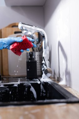 Görüntü, mavi eldiven giyen, parlak bir musluğun altında kırmızı bir sünger sıkan, sabun köpükleri ve arka planda temiz bir mutfak düzeniyle bir kişinin yakın çekimini çeker..