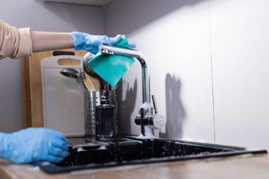 Bu görüntü, mutfak musluğunu temizlemek ve lavaboyu temizlemek için tek kullanımlık eldivenlerle bir insanı yakalıyor ve günlük yaşamdaki temizlik ve ev işlerinin önemini vurguluyor..