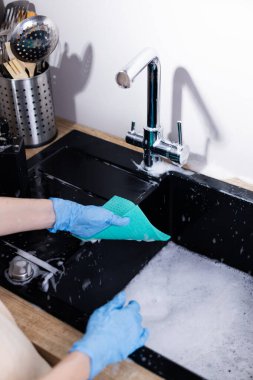 Bu resimde, mavi lastik eldivenli bir kişi, baloncuklarla dolu modern siyah bir lavaboda bulaşıkları aktif olarak yıkıyor..