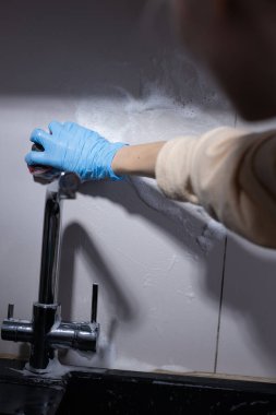 Bu resim, lavabo musluğunu ovalarken mavi eldiven takan birini yakalar, temizlik görevleri sırasında ev ortamlarında temizlik ve hijyenin önemini gösterir..