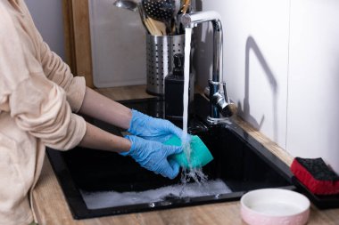 Resim, günlük mutfak işleri ve temizlik işlerinde temizliğin önemini vurgulayarak, köpüklerle dolu bir lavaboda sünger temizleyen mavi eldivenli bir kişinin resmini gösteriyor..