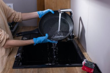 Görüntüde, tavadan lavaboya su döken mavi eldivenler giyen eller, mutfak faaliyetlerini, temizliği ve bulaşıkları özenle yıkamanın önemini vurguluyor..