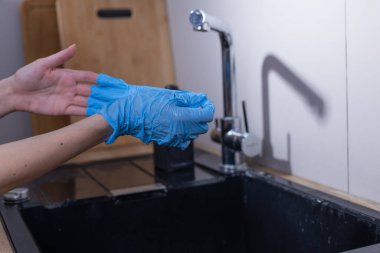 Bir çift el, parlak bir lavabo ve ahşap elementlerle çağdaş bir mutfakta temizlik görevlerine hazırlanmak için açık mavi tek kullanımlık eldivenler giyer..