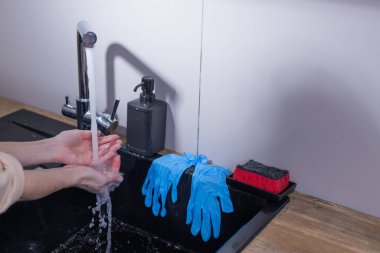 Mutfak lavabosunda akan suyun altında, ellerin yakın çekim görüntüsü, bir şişe sıvı sabun, lastik eldiven ve yakınlara yerleştirilmiş temizlik süngerleri ile birlikte..