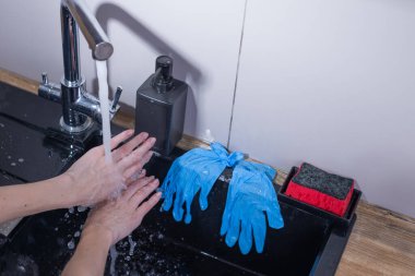 Görüntü, çalışan bir musluğun altında ellerini yıkayan bir kişinin kişisel hijyen uygulamalarını ön plana çıkarıyor. Yakınlarda, mavi eldivenler ve kırmızı bir sünger temizlik rutinini gösteriyor..